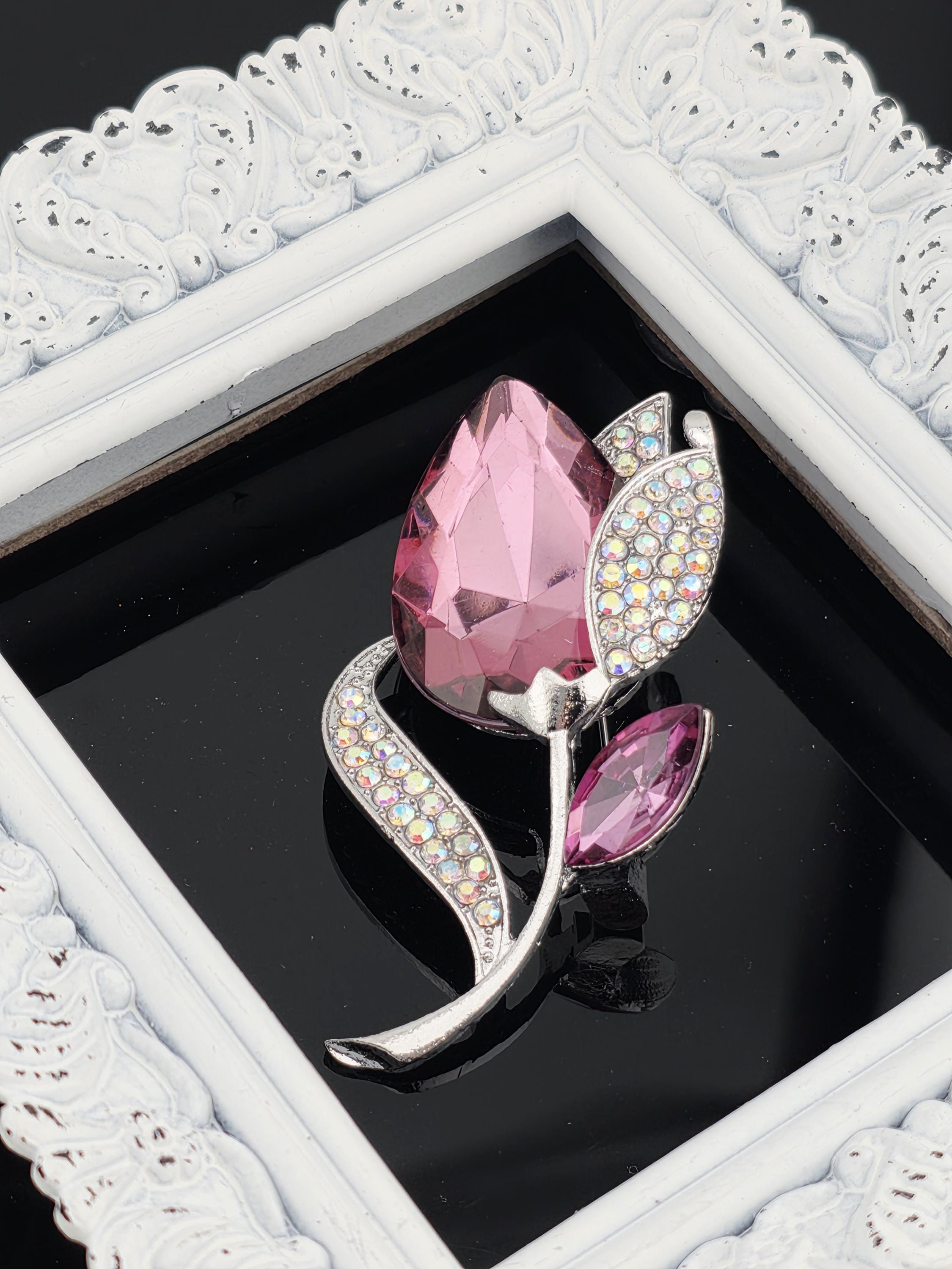 A decorative brooch with a pink gemstone and a silver leaf design, displayed in a white ornate frame.