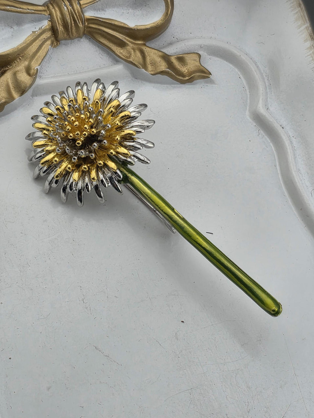 A decorative flower-shaped hair accessory with a green stem and gold and silver details, placed on a white surface.