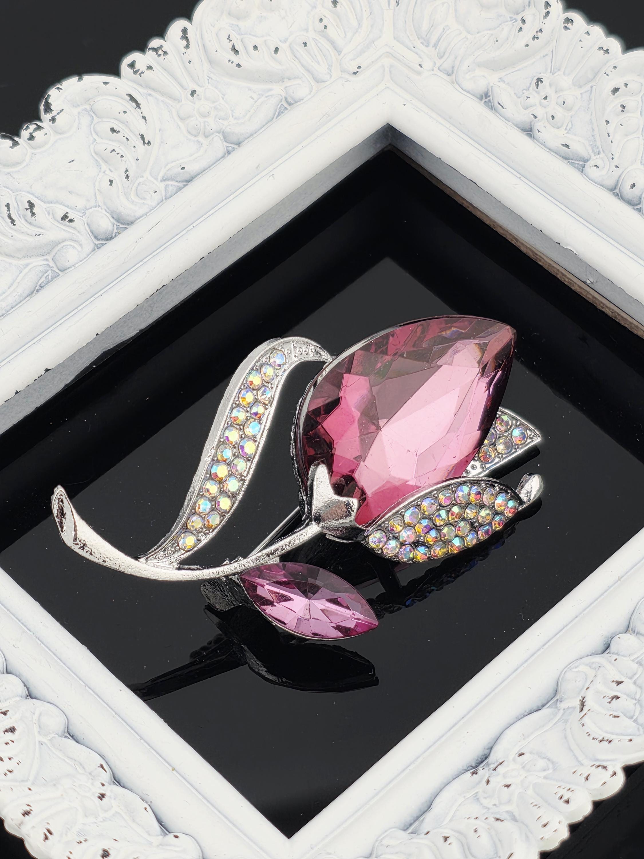 A pink crystal brooch with rhinestones is displayed in a white ornate frame against a black background.