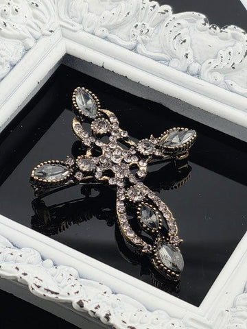 A decorative brooch with a cross design and sparkling crystal elements is displayed in a white ornate frame.