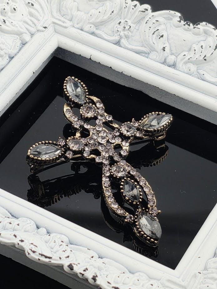A decorative brooch with a cross design and sparkling crystal elements is displayed in a white ornate frame.