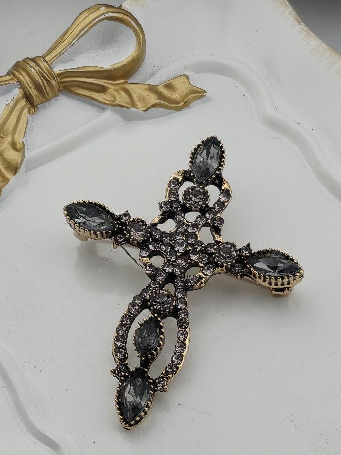 A decorative cross brooch with a gold-colored ribbon and black gemstones is displayed on a white surface.
