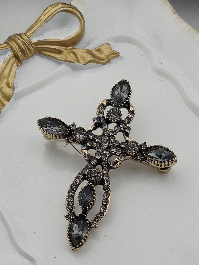 A decorative cross brooch with a gold-colored ribbon and black gemstones is displayed on a white surface.