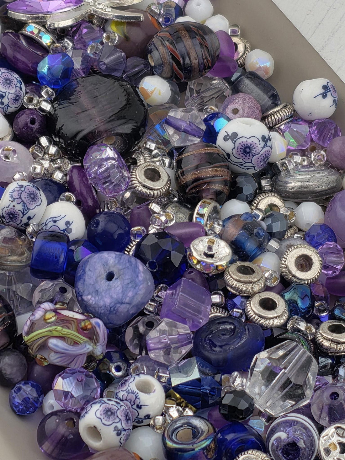 A collection of purple and blue beads of various shapes and sizes. The beads include faceted crystals, round beads with floral patterns, and dark, swirled glass beads. Silver-toned metal accents are interspersed throughout the mix..