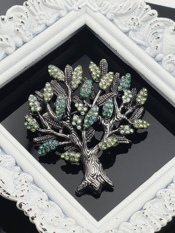 A decorative tree brooch with green and blue gemstones is displayed in a white ornate frame.