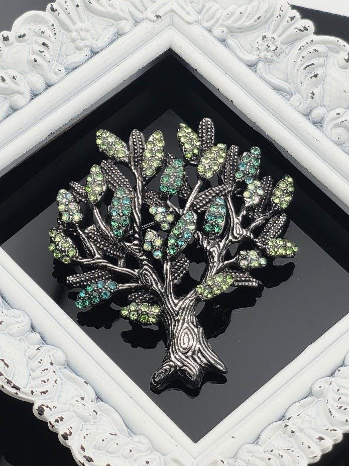 A decorative tree brooch with green and blue gemstones is displayed in a white ornate frame.