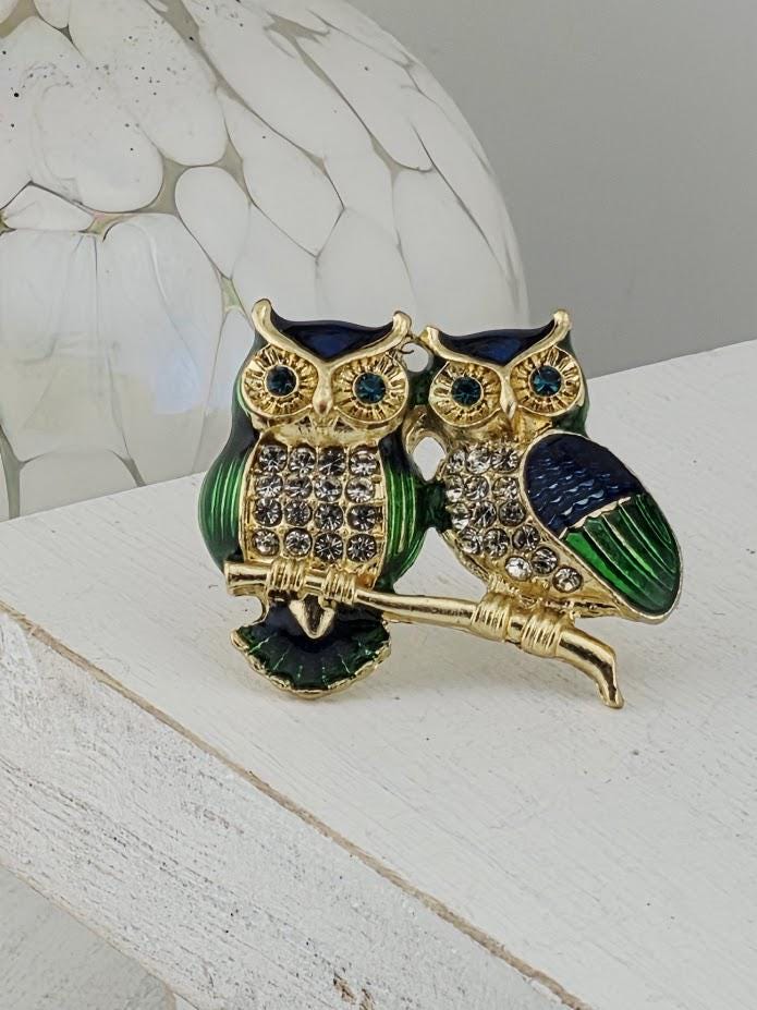 A decorative brooch featuring two ornate owls, one with green and blue accents and the other with blue and gold accents, sitting on a white surface.