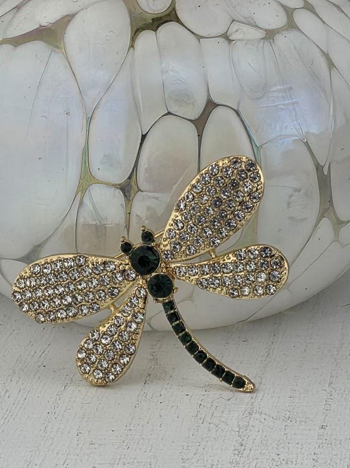A decorative brooch in the shape of a dragonfly, adorned with rhinestones and featuring a green stem.