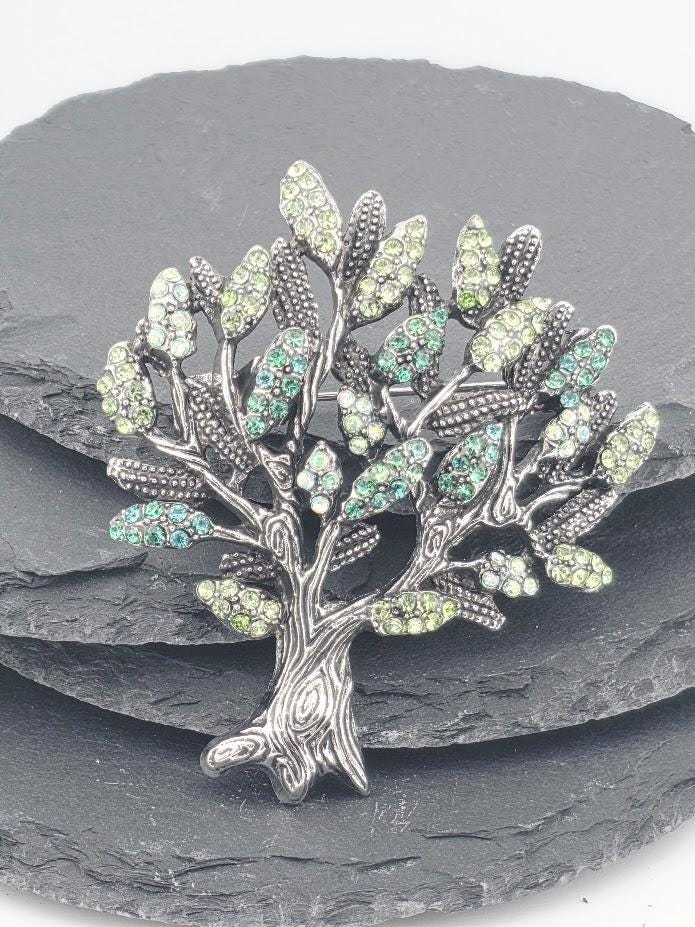 A decorative tree brooch with green and silver gemstones, placed on a gray surface.