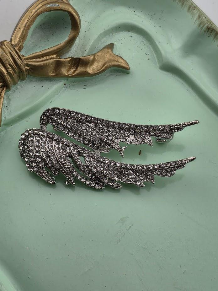 a pair of silver wing-shaped earrings with sparkling rhinestones on a light green background.