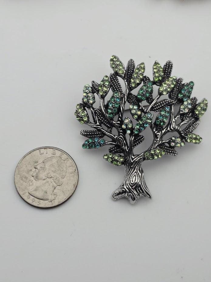 A silver-colored brooch in the shape of a tree with green and blue gemstones is placed next to a United States quarter coin.