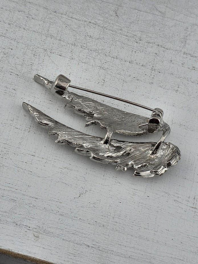 A silver pin with a bird-like design, resembling a bird's wing, is shown on a white surface.