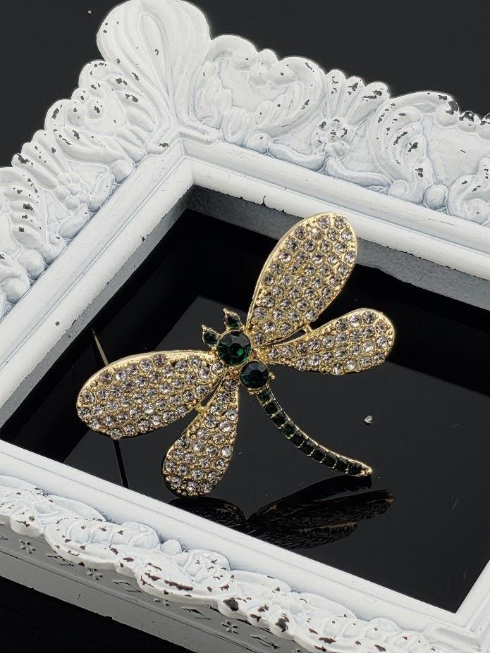 A decorative gold-colored brooch with green gemstones is displayed in a white ornate frame against a black background.
