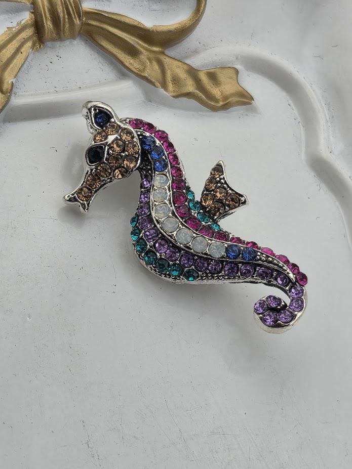 A decorative brooch in the shape of a fish, adorned with colorful gemstones, is displayed within an ornate white frame.