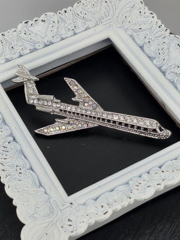 A silver airplane brooch with rhinestones is displayed in a white ornate frame.