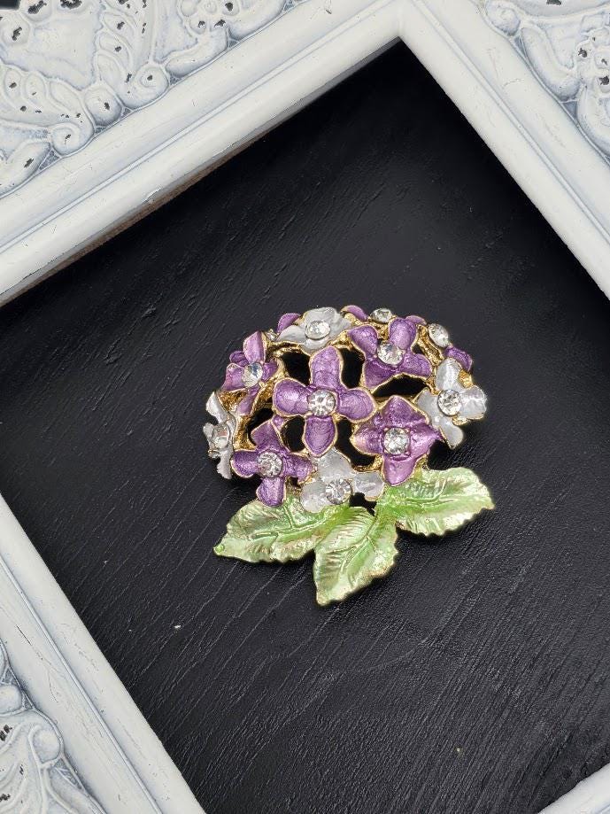 A decorative brooch with purple and white flowers and a green leaf is displayed on a black surface, framed by a white border.