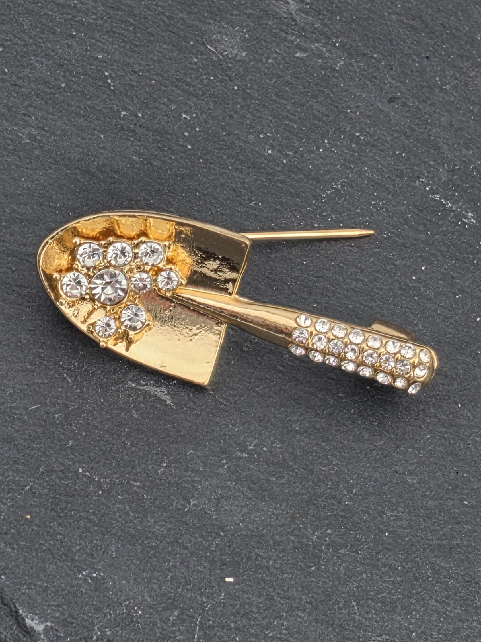A gold-colored brooch with a shovel-shaped design and numerous small diamonds or gemstones embedded in it, resting on a dark surface.