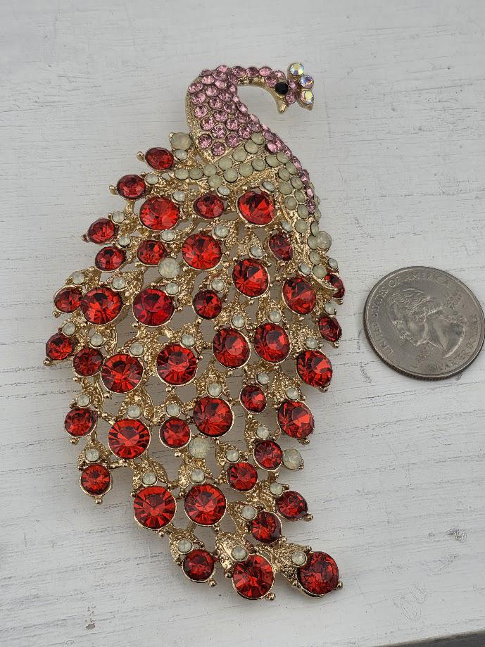 A decorative peacock brooch with a large, intricate design of red gemstones and a gold-colored head. The brooch is placed next to a coin for scale.