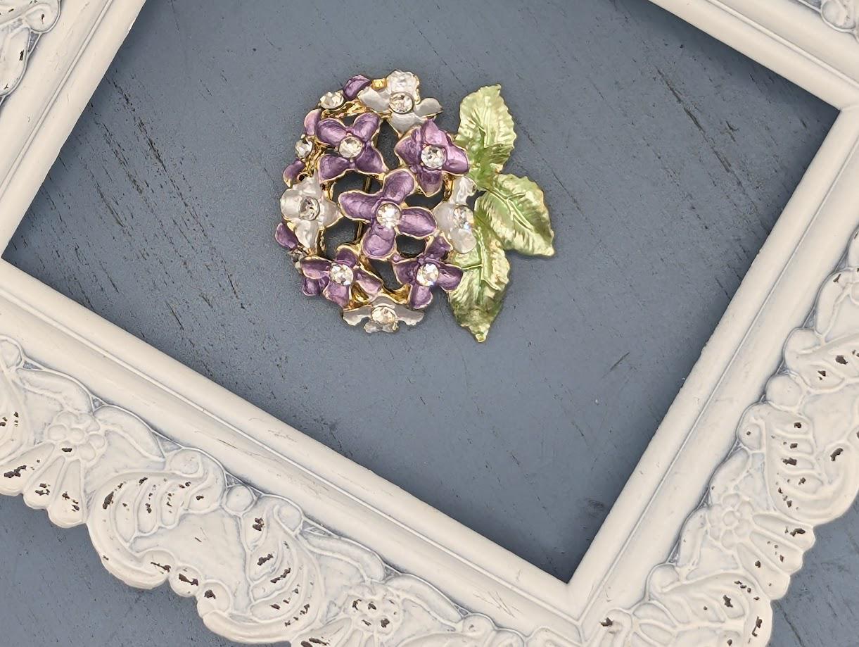 A decorative brooch with purple and green flowers is displayed in a white ornate frame against a blue background.
