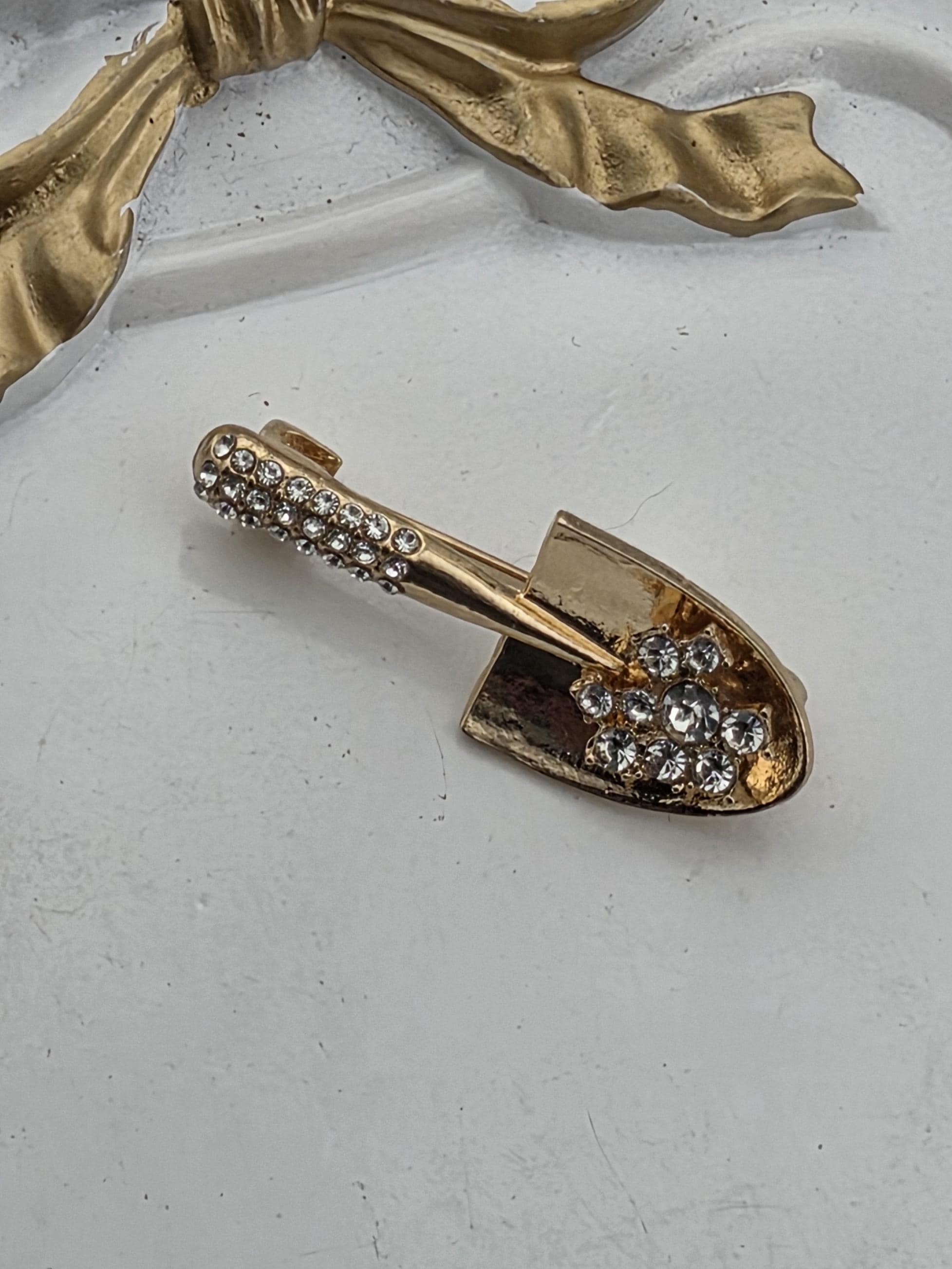 A gold-colored brooch with a diamond-encrusted shovel-shaped design is displayed against a white background.