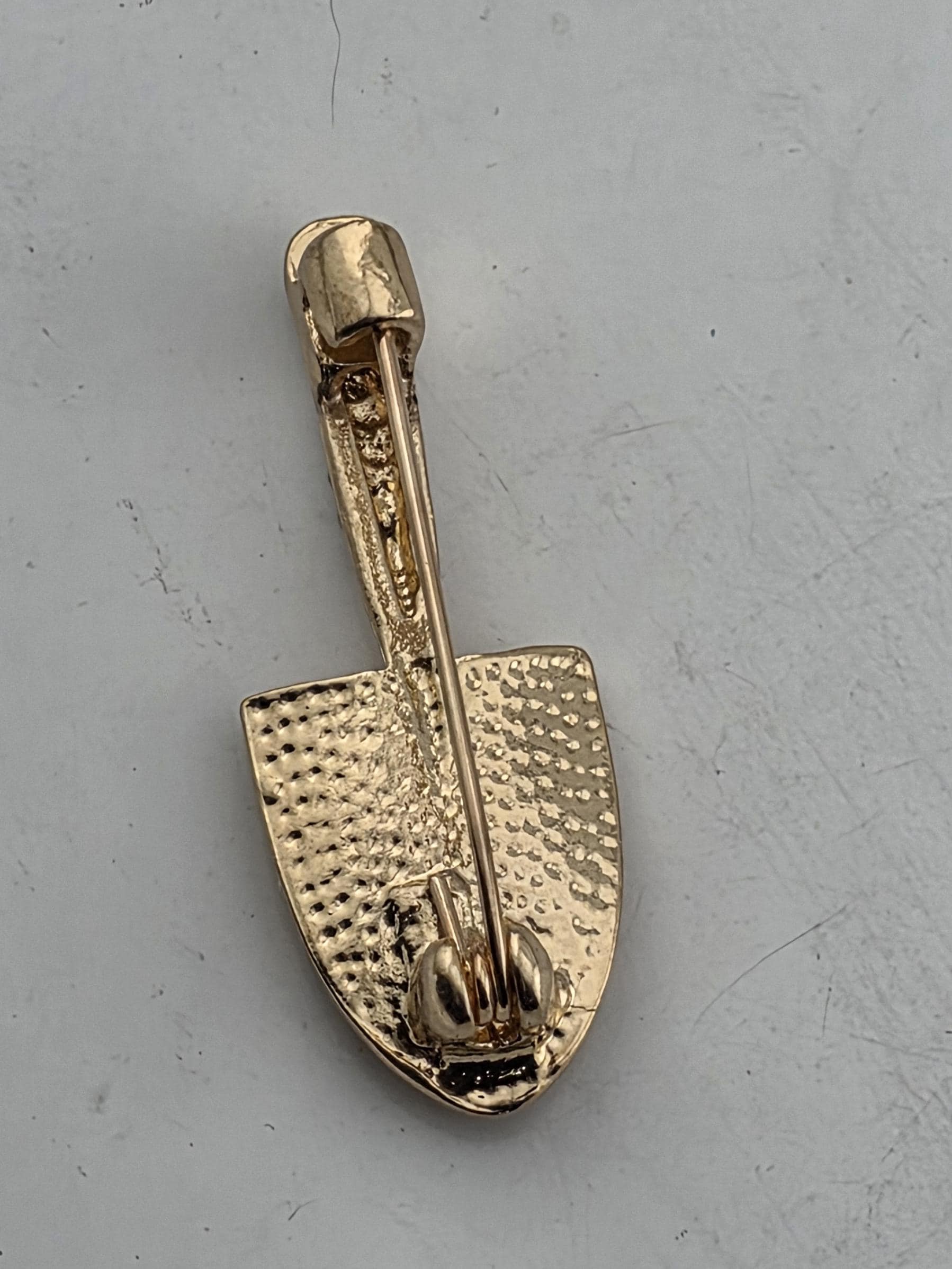 A gold-colored pin or brooch with a textured surface and a pointed end, resting on a white surface.