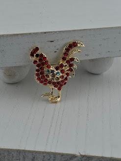 A decorative rooster brooch with red gemstones is displayed on a white surface.