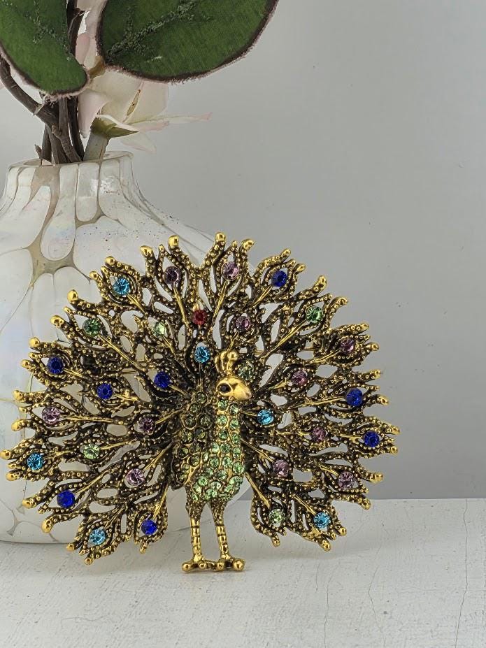 A decorative gold-colored peacock brooch with a colorful array of gemstones, including blue, green, purple, and red stones, set against a gray background.