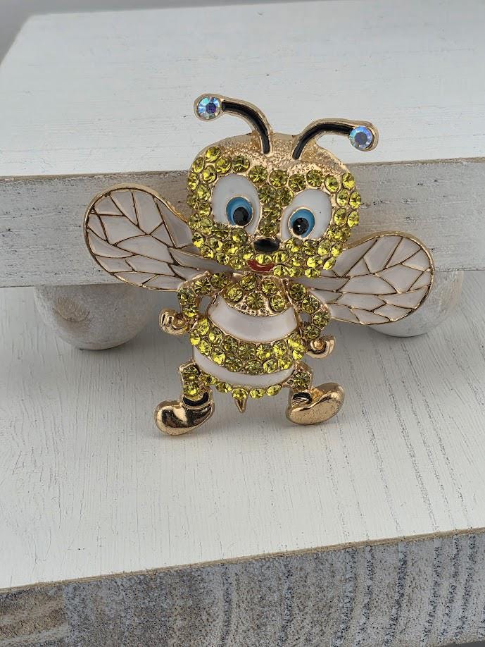 A decorative brooch in the shape of a bee, adorned with yellow and green gemstones, is displayed on a white surface.