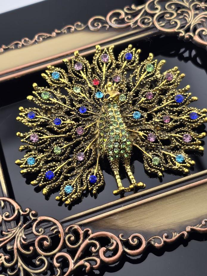 a decorative piece of jewelry, possibly a brooch or pendant, with a peacock design. The peacock is adorned with colorful gemstones and is displayed against a dark background.
