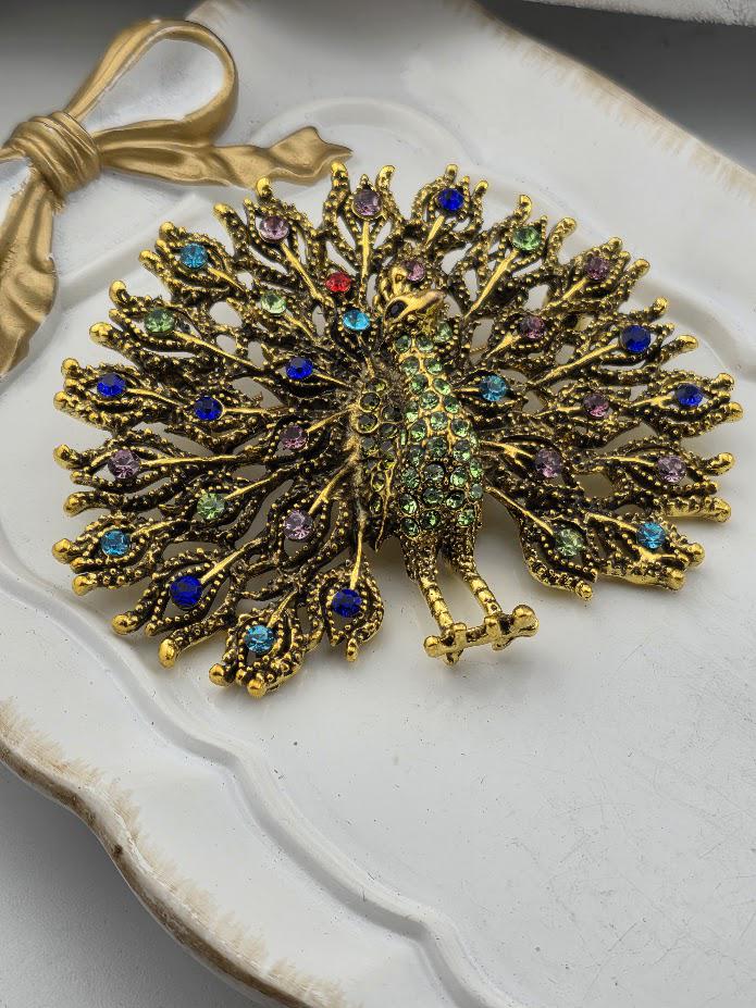 A decorative gold and colorful peacock brooch with a detailed design and sparkling gemstones.
