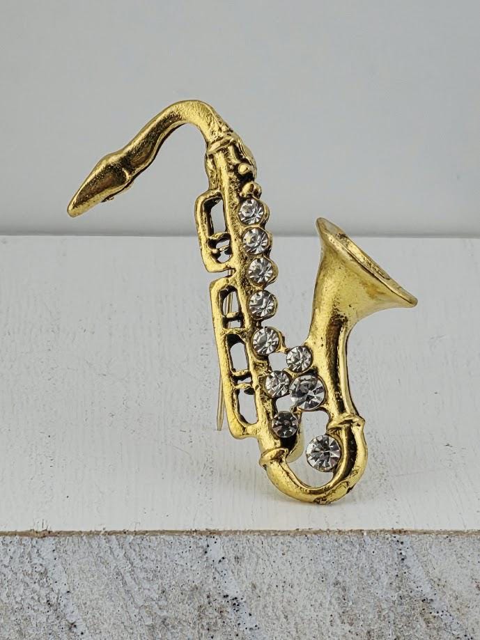 A gold-colored saxophone brooch with rhinestones on it, placed on a white surface.