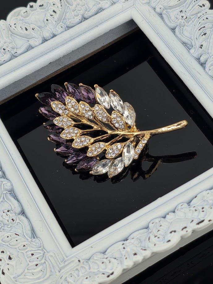 A decorative brooch in the shape of a leaf, with a gold frame and a combination of purple and white gemstones.