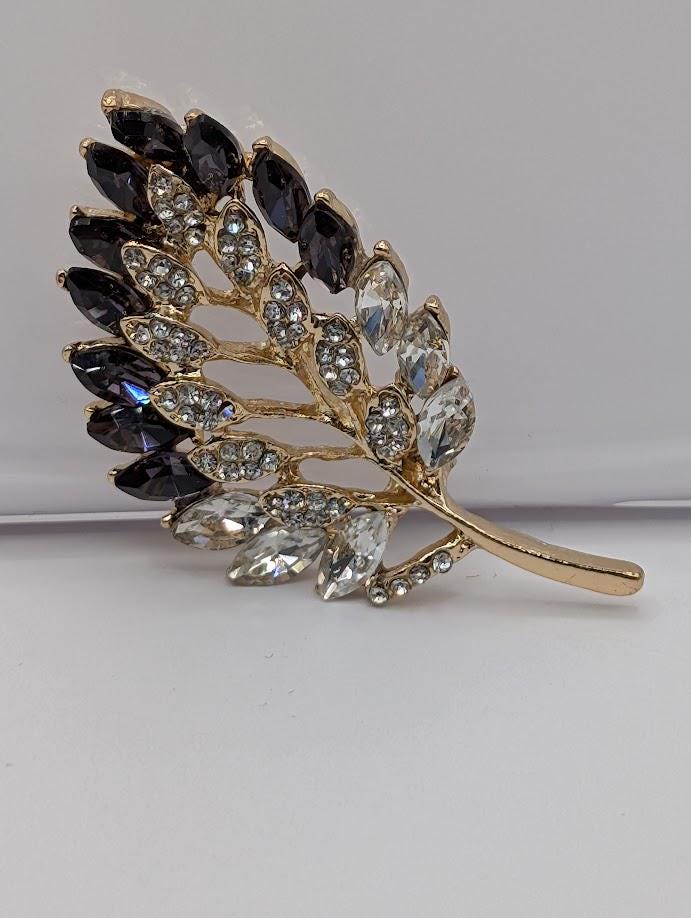 A decorative leaf-shaped brooch with a gold-colored stem and a branch adorned with black and clear gemstones.