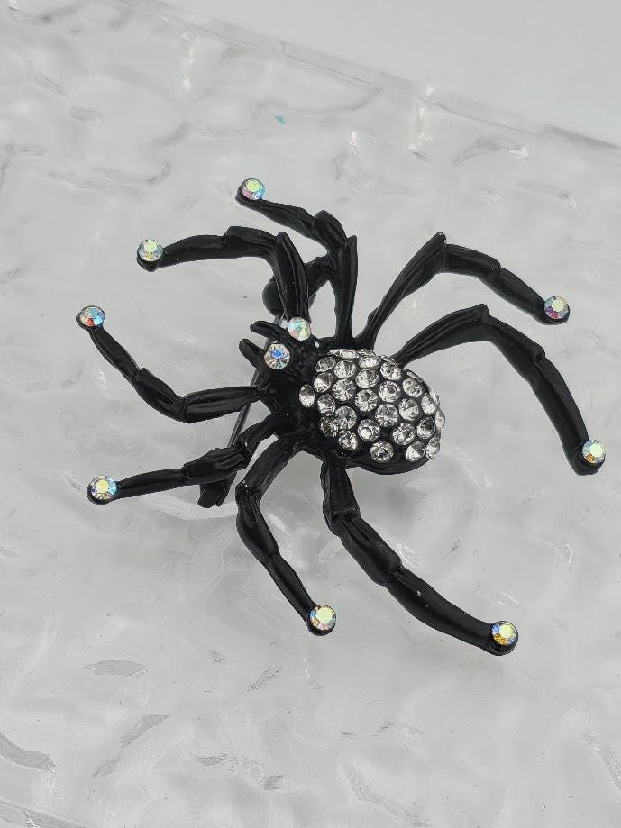 A black spider-shaped piece of jewelry with rhinestones or crystals on it, set against a white background.