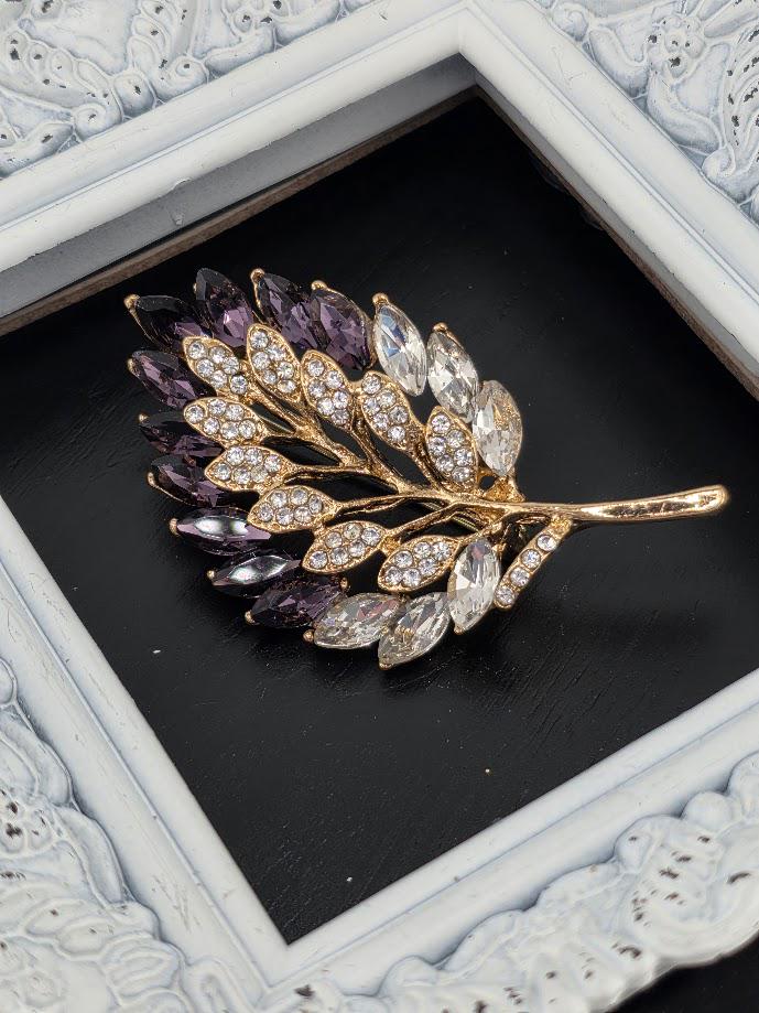 A decorative brooch in the shape of a leaf, with a gold frame and a branch adorned with purple and clear gemstones.