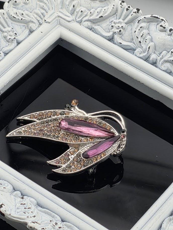 A silver brooch with a butterfly design, adorned with pink and brown gemstones, is displayed in a white ornate frame.