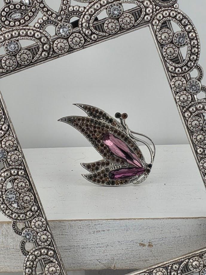 A silver-colored ornate frame with a butterfly-shaped design and a purple gemstone on it, placed on a white surface.