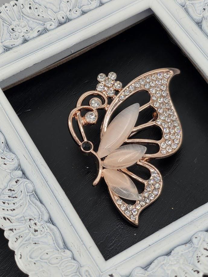 A decorative brooch in the shape of a butterfly, adorned with rhinestones, is displayed within a white ornate frame.