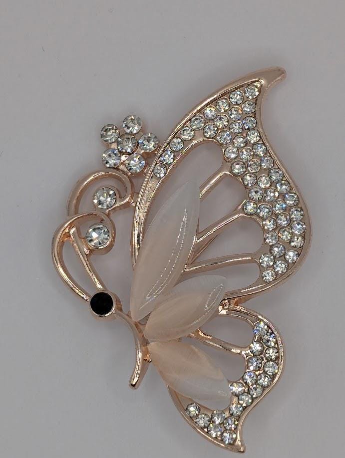 A decorative butterfly brooch with a gold-colored metal frame and a beige-colored butterfly wing adorned with small, clear gemstones.