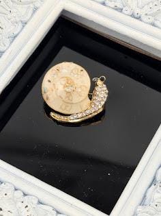 A gold-colored brooch in the shape of a snail, adorned with small diamonds, is displayed on a black surface within a white frame.
