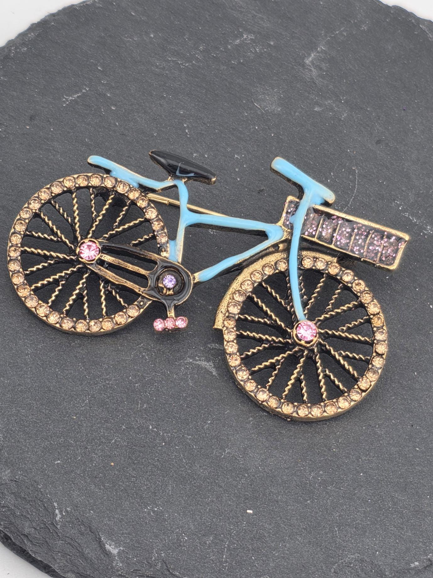 A decorative bicycle brooch with a blue frame and gold accents, including rhinestones and a pink gemstone on the handlebars.
