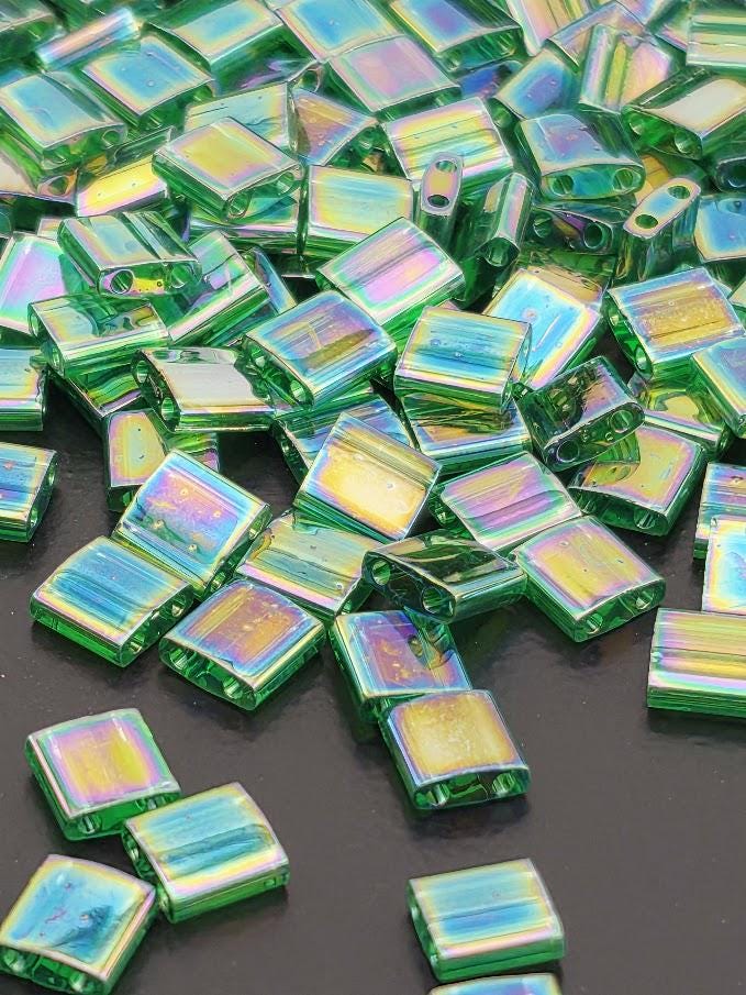 The image is a close-up of numerous green glass beads scattered on a dark surface. The beads are of various shapes and sizes, creating a visually interesting and colorful display.