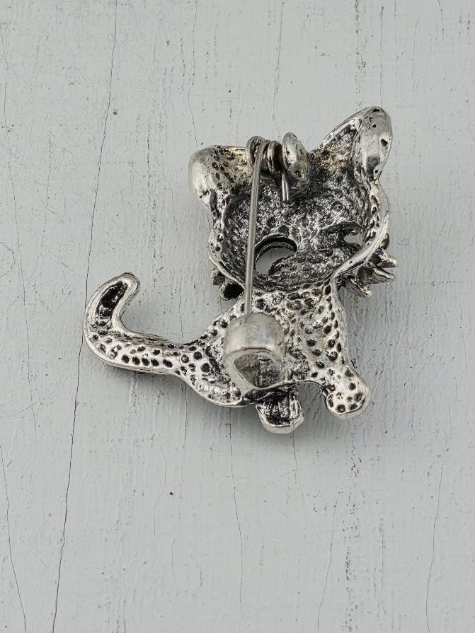A silver-colored metal pin or brooch in the shape of a cat, with a pointed nose and a small eye, is shown against a white background.