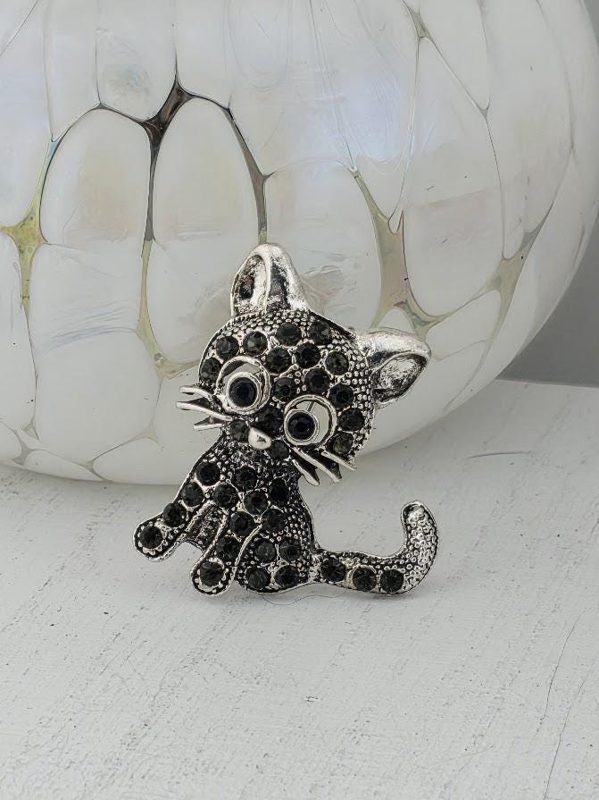 A silver brooch in the shape of a cat is displayed on a white surface, with a white and gold vase in the background.