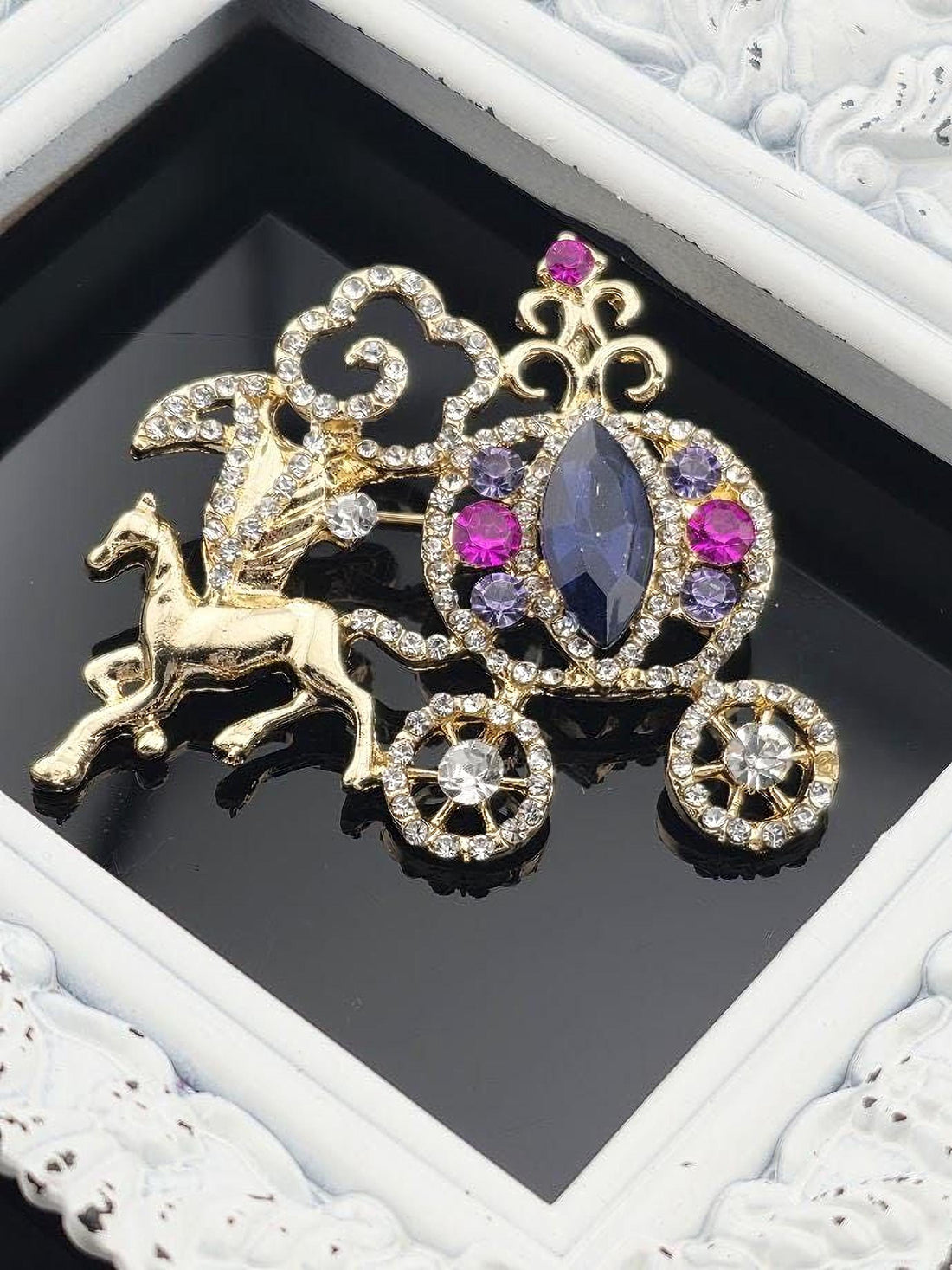 A decorative brooch with a gold horse and carriage motif, adorned with gemstones and a large blue gemstone.