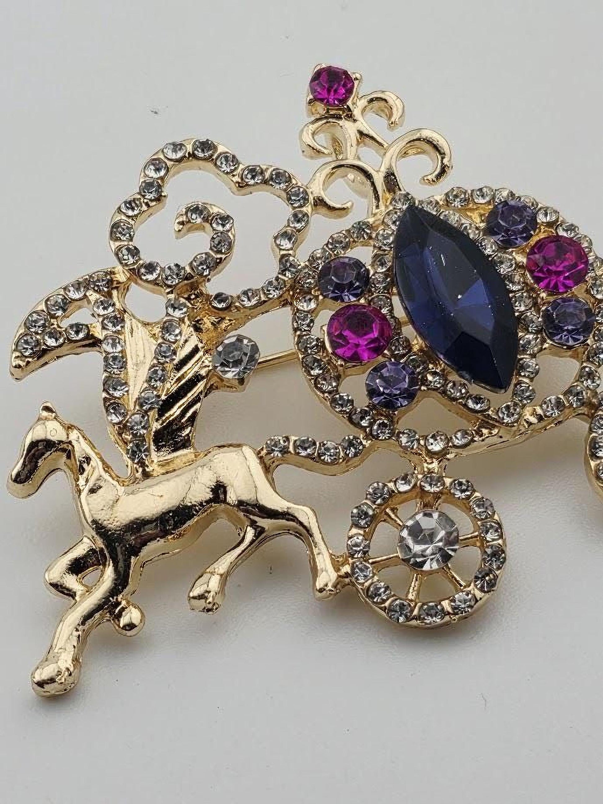 A decorative brooch with a gold-colored horse design and various gemstones, including purple and blue stones, as well as a large central gemstone.