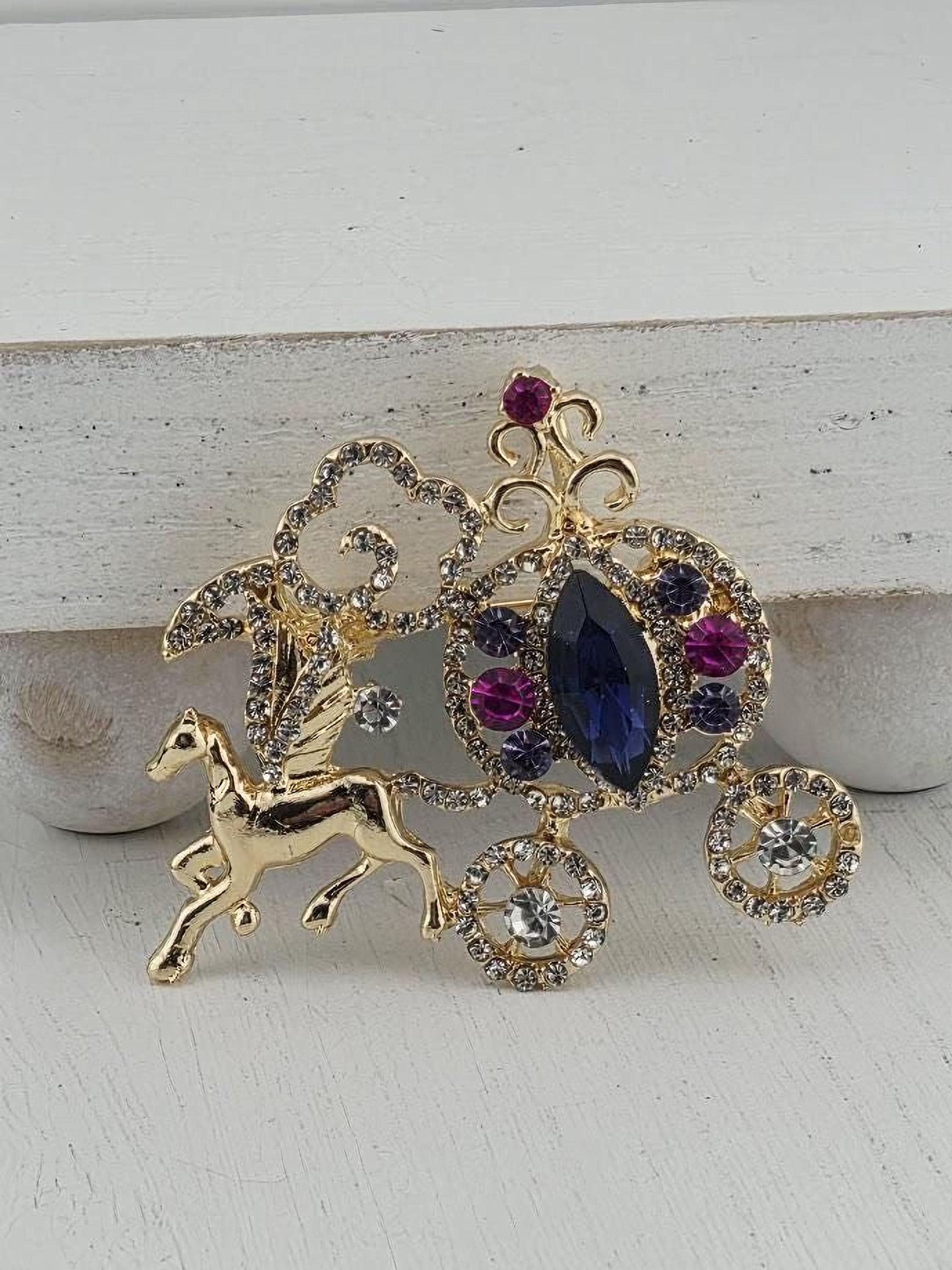 A decorative brooch with a gold horse and carriage motif, adorned with gemstones and a large blue gemstone.