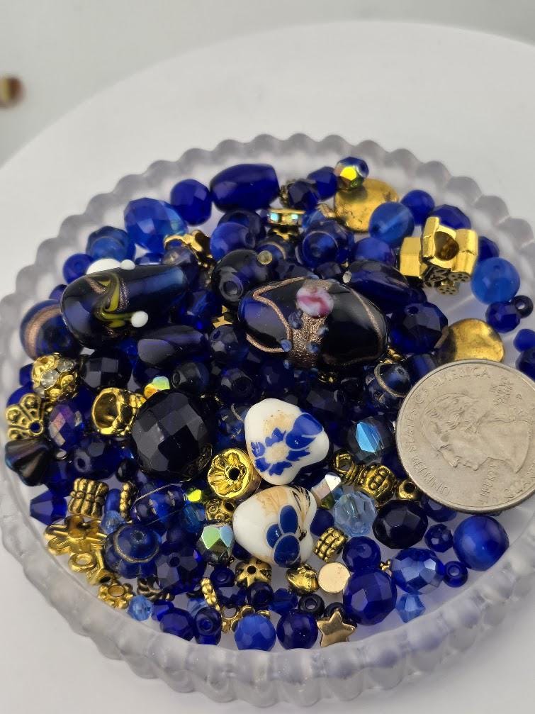 A collection of various blue and gold beads, including glass beads, metal beads, and other decorative elements, arranged in a glass container.