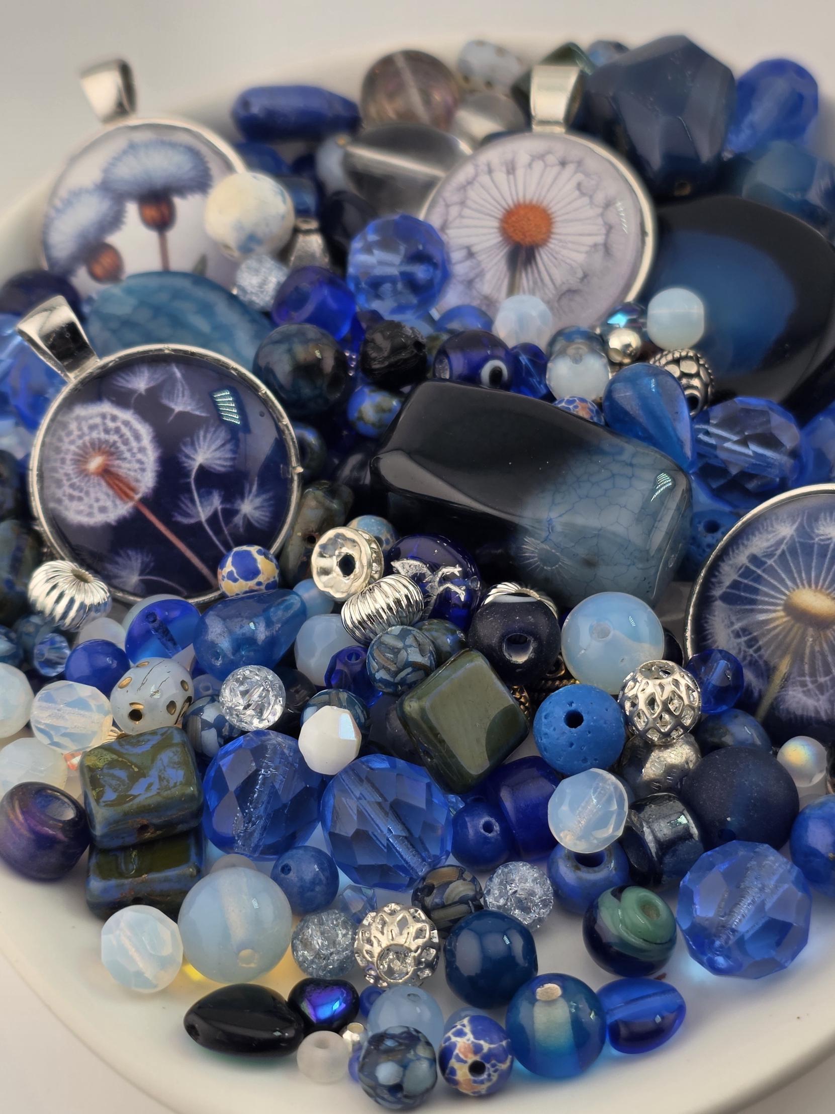 The image is a close-up of a collection of various blue and silver beads, charms, and other decorative items arranged on a white surface.