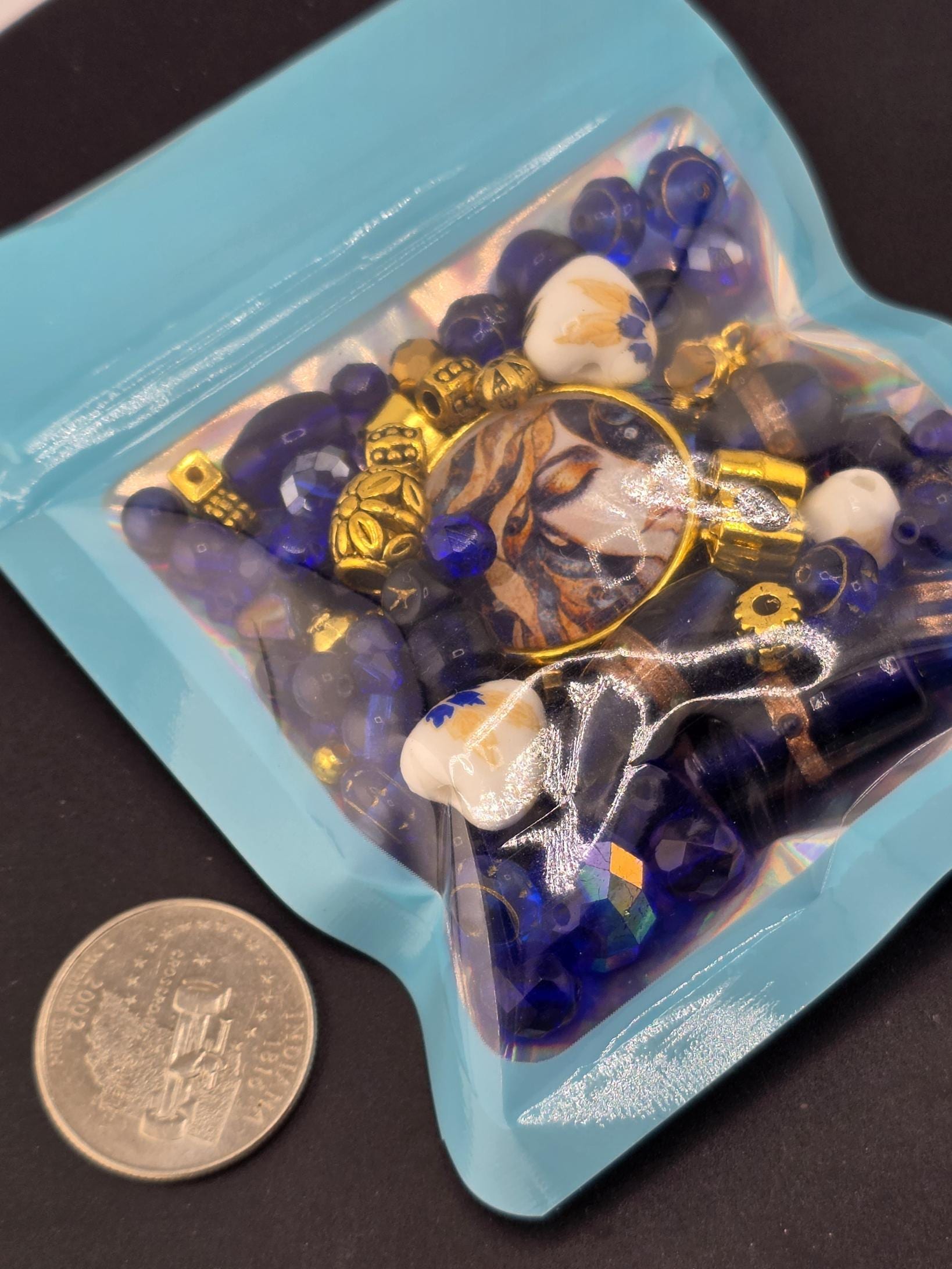 A blue bag filled with various colorful beads and a gold-colored pendant is placed next to a coin.